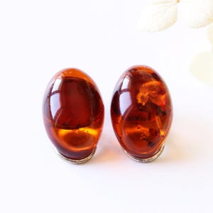 May include: Two oval amber earrings with a silver base are shown against a white backdrop. The earrings have a glossy, translucent finish, highlighting the rich, warm hues of the amber. The letter "A" is in the bottom right corner.