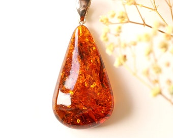 May include: A teardrop-shaped amber pendant with a silver bail. The amber is a deep orange color with inclusions of plant matter.