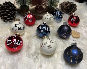 May include: A collection of personalized Christmas ornaments. The ornaments are round, with a glossy finish in various colors including red, white, and blue. Each ornament features a name in elegant script and a snowflake design.