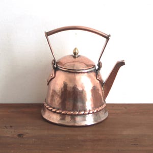 Op de afbeelding: Een koperen ketel met een gehamerde afwerking, met een gebogen handvat en een tuit. De ketel heeft een decoratief touwachtig detail rond de basis en een kleine messing knop op het deksel. De ketel wordt op een houten oppervlak gepresenteerd.
