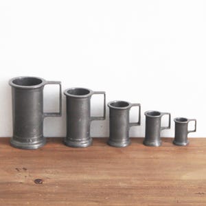 May include: A set of five antique pewter tankards of different sizes. Each tankard has a cylindrical body, a flat base, and a rectangular handle. The tankards are arranged in a row on a wooden surface, set against a white backdrop.