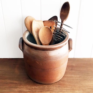 May include: A brown ceramic crock filled with wooden kitchen utensils, including spoons, a spatula, and a whisk. The crock has two handles and sits on a wooden surface against a white background. The utensils are various shades of light brown.