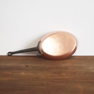 Puede incluir: Una sartén de cobre con un mango oscuro y remaches. La sartén tiene una superficie pulida y reflectante y está colocada sobre un fondo blanco y una superficie de madera.