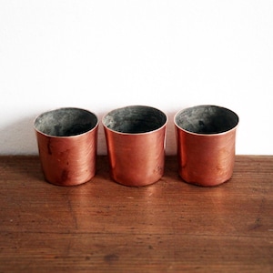 May include: Three copper-coloured cylindrical containers with a dark interior are arranged on a wooden surface. The containers have a smooth, reflective finish and a slightly tapered shape. The background is a plain white wall.