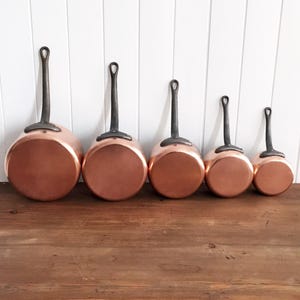 May include: A set of five copper saucepans with dark metal handles. The pans are arranged in a row, decreasing in size from left to right. The copper surfaces reflect light, and the handles have a loop for hanging. The pans are on a wooden surface.