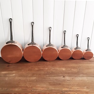 May include: A collection of six copper saucepans with long, dark metal handles. The pans are arranged in a row, decreasing in size from left to right. The copper bodies have a shiny, reflective surface, and the handles have a loop at the end.