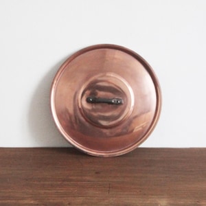 May include: A copper pot lid with a dark handle. The lid is round with a slightly raised center and a polished finish. The copper reflects light, creating a warm, inviting aesthetic. The lid is sitting on a wooden surface.