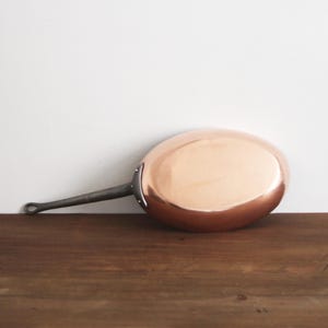 May include: A copper frying pan with a long, dark metal handle. The pan is oval-shaped and rests on a wooden surface. The handle has a loop at the end for hanging. The pan is a kitchenware item.