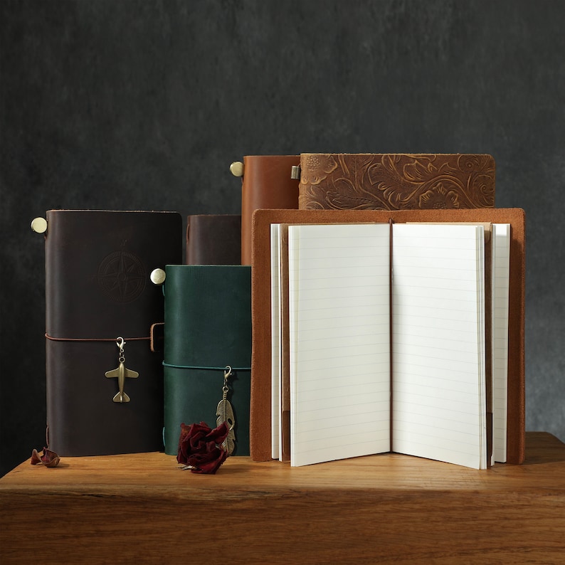 May include: Four leather-bound journals with decorative accents. The journals are stacked on a wooden surface. The top journal is open, revealing blank lined pages.