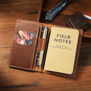May include: A brown leather notebook cover featuring multiple interior card slots, a central pen loop holding a silver pen, and a Field Notes 48-page memo book. The cover is open, displaying two credit cards in the slots and a passport tucked behind the pen.