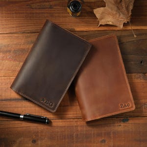 May include: Two rectangular leather notebook covers in dark brown and light brown. The dark brown cover features the embossed text "BG" with a star symbol, while the light brown cover is embossed with the name "Zac" in the bottom right corner.