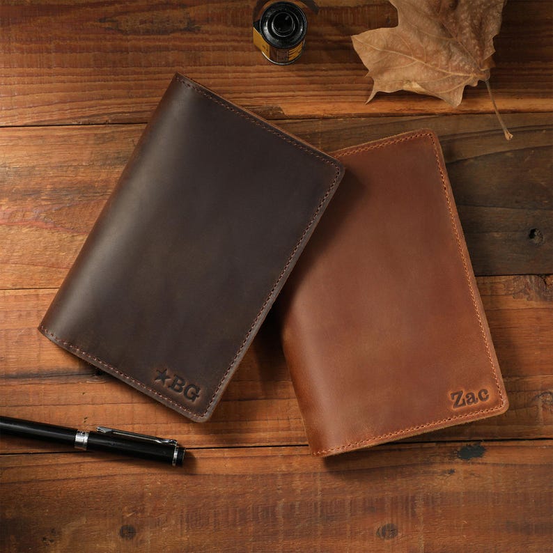 May include: Two rectangular leather notebook covers in dark brown and light brown. The dark brown cover features the embossed text "BG" with a star symbol, while the light brown cover is embossed with the name "Zac" in the bottom right corner.