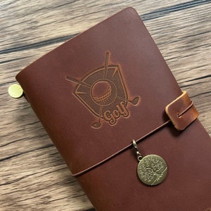May include: Brown leather journal with a golf ball and clubs design embossed on the front cover. The journal has a brown leather strap with a metal tree charm attached.