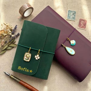 May include: A dark green leather journal with the name Sofia and a star printed in gold, featuring two gold-toned charms of a flower and a clover. Beside it is a dark purple journal with two gold-toned charms, a fountain pen, dried flowers, and stamps.