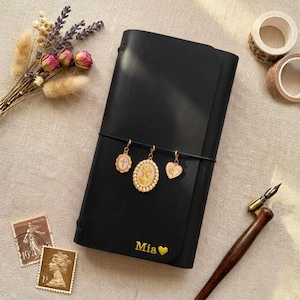 May include: A black leather journal featuring the name Mia and a heart symbol in gold lettering. Three gold-toned charms, including a cross, a cameo portrait, and a heart, are attached to a black elastic band wrapped around the center of the cover.