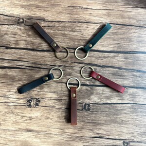May include: Five leather keychains with metal rings. The keychains are in different colors: brown, dark green, black, burgundy, and a lighter brown. The keychains are arranged in a star shape on a wooden surface.