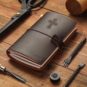 May include: A dark brown leather journal with a wrap-around strap closure. The cover features an embossed cross symbol and the name Ruth. The journal contains multiple white paper inserts and is surrounded by various metal crafting tools.
