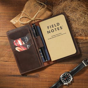 May include: A dark brown leather notebook cover featuring multiple card slots, a pen loop with a black pen, and a Field Notes memo book with tan covers. The notebook cover holds a blue passport and two credit cards, while a wristwatch sits nearby.