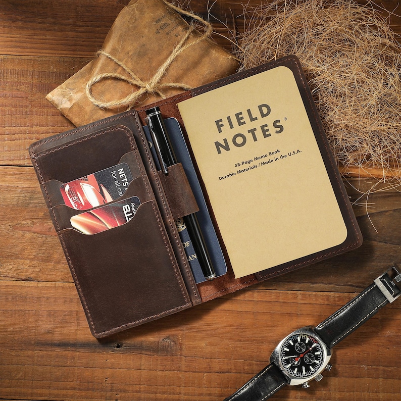 May include: A dark brown leather notebook cover featuring multiple card slots, a pen loop with a black pen, and a Field Notes memo book with tan covers. The notebook cover holds a blue passport and two credit cards, while a wristwatch sits nearby.