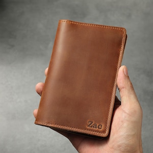 May include: A vertical brown leather wallet featuring a smooth, matte finish and visible stitching along the edges. The bottom right corner of the wallet is embossed with the name Zac in a clear, dark, sans-serif font, showcasing a simple design.