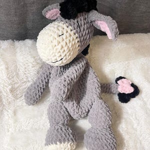 May include: A handmade crochet donkey plush toy. The donkey is primarily gray with a white face and belly, black mane, and pink inner ears. The plush animal has a smiling face and is sitting on a white, fluffy surface.