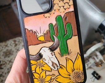 May include: A black leather phone case with a western desert scene. The case features a sunset, a cactus, a cow skull, and sunflowers.