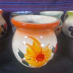 May include: A white ceramic mug with an orange rim and a hand-painted yellow flower with a red center. The mug has green leaves and a white background.