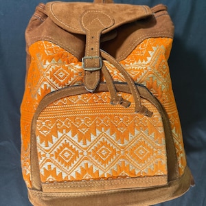 May include: A brown and orange backpack with a flap closure and a front pocket. The backpack features a geometric pattern in orange and gold, with brown suede accents and a buckle closure. The bag has a drawstring closure and adjustable straps.