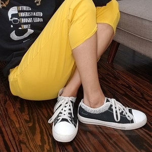 May include: A pair of black canvas sneakers with white soles and laces. The sneakers have a white crocheted trim around the ankle and the word "I dissent" written on the side. The person wearing the sneakers is sitting on a wooden floor wearing yellow pants.
