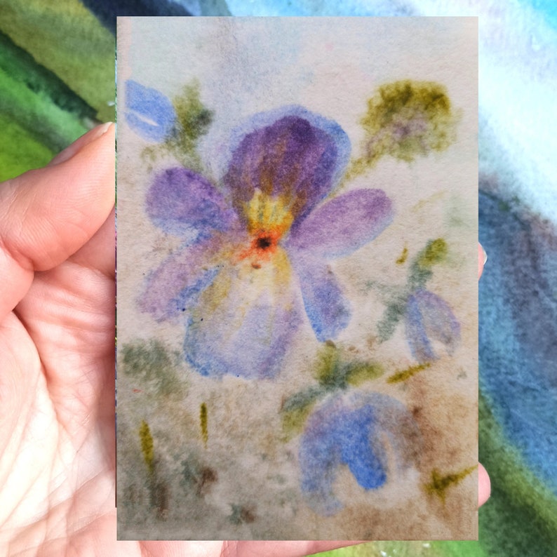 May include: Watercolor painting of a purple flower with a yellow center, surrounded by green leaves and other blue flowers on a light brown background.