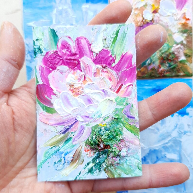 May include: A miniature oil painting of a peony flower. The artwork features thick brushstrokes in shades of pink, white, and green, creating a textured effect. The painting is held in a hand, showcasing the small scale of the piece.