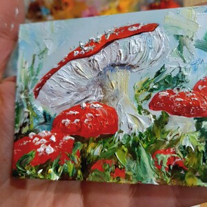 May include: An oil painting on a small rectangular canvas featuring several red mushrooms with white spots and thick, textured white gills. The mushrooms are nestled among vibrant green foliage against a pale blue sky background.