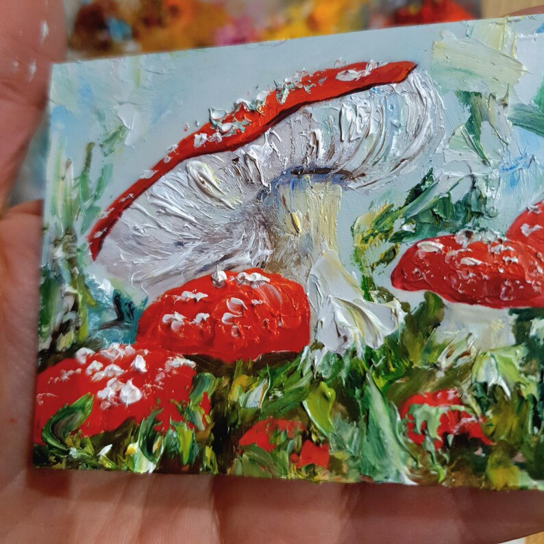 May include: An oil painting on a small rectangular canvas featuring several red mushrooms with white spots and thick, textured white gills. The mushrooms are nestled among vibrant green foliage against a pale blue sky background.