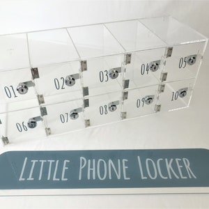 May include: A clear acrylic phone locker with ten individual compartments, each with a lock and a number label. The locker has a blue sign that reads "LITTLE PHONE LOCKER".