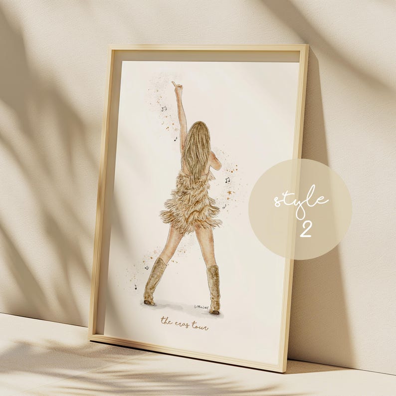 May include: Watercolor print of a performer on stage, wearing a fringed dress and boots, with an arm raised. The artwork is framed in a light wood frame. The text "the eras tour" is at the bottom of the image.