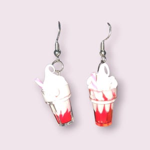 May include: A pair of novelty earrings featuring miniature ice cream floats. Each earring has a silver-tone fish hook and a clear plastic cup filled with red liquid, topped with white whipped cream and a pink straw.