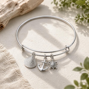 May include: A silver-colored bangle bracelet with three charms: a seashell, an anchor, and a turtle. The bracelet is adjustable and has a simple, elegant design. The charms are detailed and add a nautical theme.