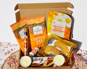 May include: A brown cardboard box filled with various self-care items, including a face mask, eye masks, coffee bags, chocolate, and candles. The box is sitting on a wooden surface with pink flower petals scattered around it.
