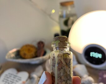 May include: A small glass bottle with a cork stopper filled with dried herbs and sealed with green wax. The bottle is being held by a hand with white nail polish.