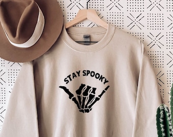 Może przedstawiać: Beżowa bluza z napisem "STAY SPOOKY" nad czarnym gestem dłoni szkieletu. Brązowy kapelusz z białym paskiem wisi po lewej stronie. Po prawej stronie znajduje się kaktus.