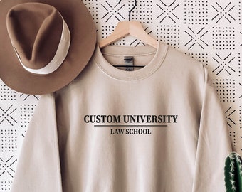 Puede incluir: Una sudadera de color beige claro con el texto "CUSTOM UNIVERSITY LAW SCHOOL" impreso en la parte delantera.
