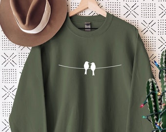 May include: A green sweatshirt with a white outline of two birds perched on a wire. The sweatshirt is hanging on a hanger against a white background with a black and white geometric pattern.