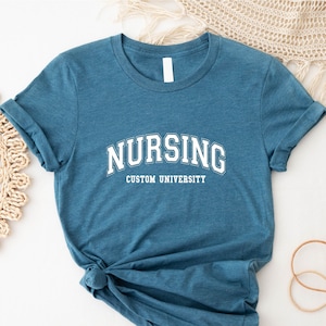 May include: Teal-colored t-shirt with the word "NURSING" in a white, arched font above "CUSTOM UNIVERSITY". The shirt has rolled-up sleeves and a knot at the bottom. The shirt is made of a soft material.