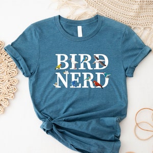 May include: A teal blue t-shirt with the words "BIRD NERD" in white letters. The words are surrounded by colourful bird illustrations.
