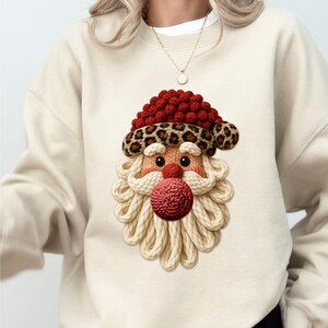 Peut inclure: Sweat-shirt beige avec un motif de Père Noël. Le visage du Père Noël a un chapeau rouge avec un bord léopard, une barbe tricotée et un nez rouge. Le motif est centré sur le devant du sweat-shirt.