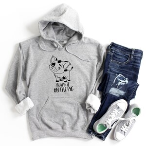 Puede incluir: Una sudadera gris con capucha con un cerdo de dibujos animados en blanco y negro y el texto "Blame it on the pig". La sudadera se combina con jeans azules y zapatillas grises.