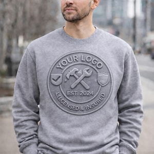 May include: A gray sweatshirt with a circular logo featuring a hammer, wrench, and shield. The logo includes the text "YOUR LOGO", "EST. 2024", and "LICENSED & INSURED". The sweatshirt is a crew neck style.