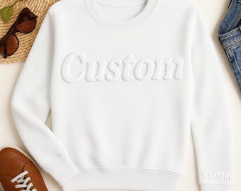 May include: A white sweatshirt with the word "Custom" embossed across the chest. The word "Custom" is also embroidered on the cuff of the left sleeve. The sweatshirt is displayed with brown shoes, sunglasses, and a straw hat.