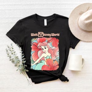 A black t-shirt with a Disney-themed graphic of a mermaid, a white mug, a beige hat, and some greenery on a white background.
