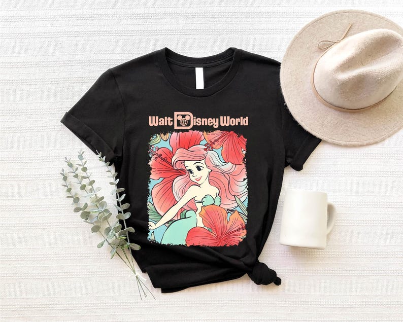 A black t-shirt with a Disney-themed graphic of a mermaid, a white mug, a beige hat, and some greenery on a white background.